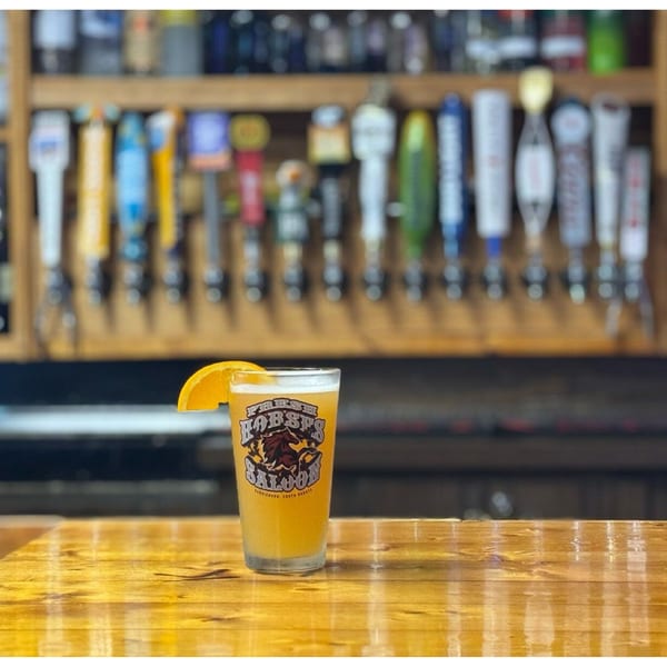 A Fresh Horses Saloon beer glass with an orange slice sits on a wooden bar, with tap handles in the background.