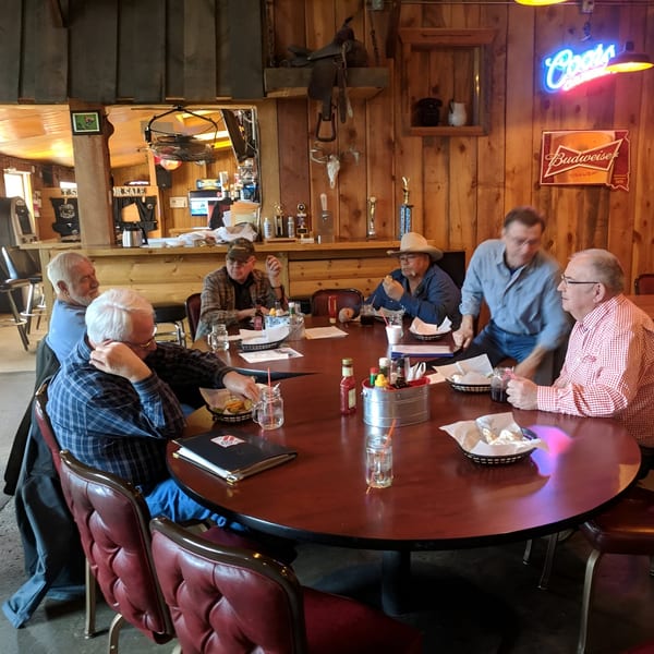 Men gathered around a large round table at Fresh Horses Saloon, with rustic wood paneling and neon signs.